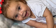 Lola is registered to the contest to win money with this photo: baby, child, smiling, happy, face, eyes, hair, bows, white_clothing, blanket, pattern, infant, lying_down, cute, skin, arm, hands, indoors, portrait, young