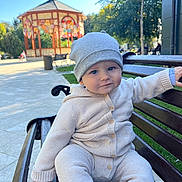 Joshua is registered to the contest to win money with this photo: baby, child, bench, park, outdoor, knitwear, beanie, cute, smiling, person, daylight, gazebo, trees, greenery, public_space, wood, sitting, face, blue_eyes, casual