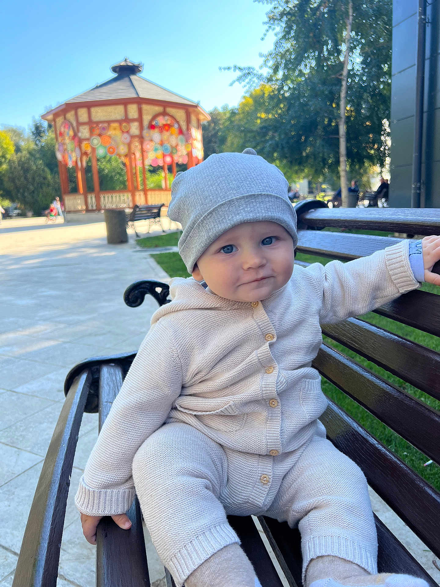 Joshua is registered to the contest to win money with this photo: baby, child, bench, park, outdoor, knitwear, beanie, cute, smiling, person, daylight, gazebo, trees, greenery, public_space, wood, sitting, face, blue_eyes, casual