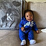 baby, smiling, blue_outfit, couch, blanket, embroidery, letter_k, happy, indoor, child, sitting, sock, curly_hair, person, furniture, home, cozy, fabric, textile, portrait