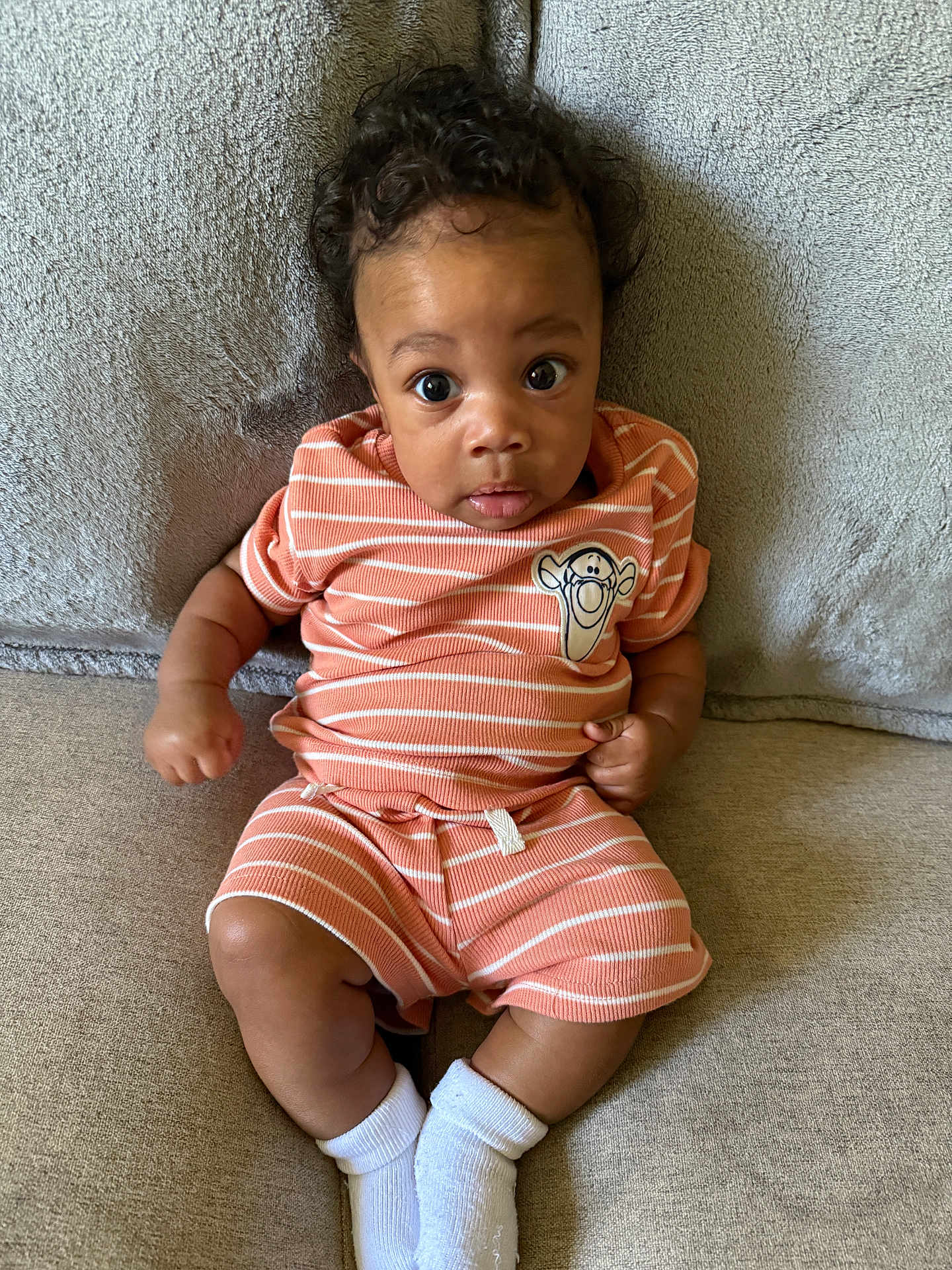 Joshua joined the competition — help win amazing prizes! baby, infant, child, couch, clothing, striped_outfit, socks, face, curly_hair, expression, indoor, portrait, cute, small_hands, seated, soft_texture, cartoon_patch, orange, white, cozy