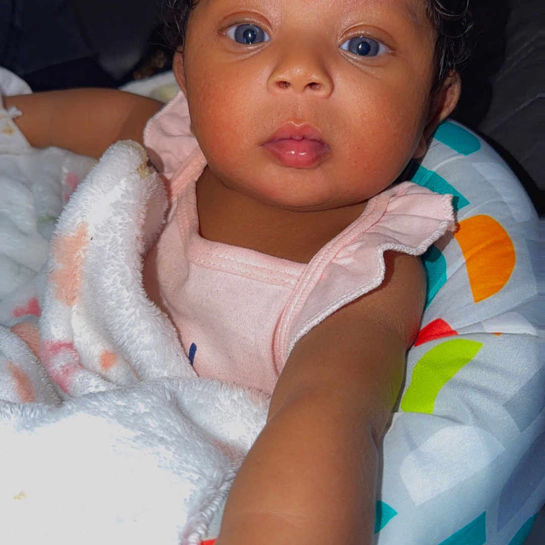 Jahlani joined the competition — help win amazing prizes! baby, infant, child, blue_eyes, blanket, baby_chair, curly_hair, reaching_hand, pink_clothing, indoor, portrait, cute, face, skin, person, young, seated, colorful, soft_light, cozy