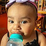 Xiomara is registered to the contest to win money with this photo: toddler, baby, child, bottle, headband, purple_bow, black_dress, indoor, closeup, face, eyes, curious, sitting, furniture, wooden_chair, drink, feeding, person, cute, young_child