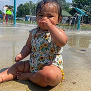 Xiomara joined the competition — help win amazing prizes! toddler, child, water_park, splash_pad, water_spray, sunlight, swimsuit, wet_skin, outdoor, summer, playground, concrete, greenery, trees, people, fun, sunflower_pattern, sitting, cute, water