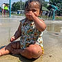 toddler, child, water_park, splash_pad, water_spray, sunlight, swimsuit, wet_skin, outdoor, summer, playground, concrete, greenery, trees, people, fun, sunflower_pattern, sitting, cute, water