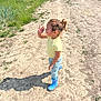 Willow-Rose is registered to the contest to win money with this photo: child, toddler, outdoor, dirt_path, grass, sunlight, boots, yellow_shirt, blue_boots, waving, curly_hair, pony_tail, nature, playful, portrait, casual_clothing, daytime, shadow, smiling, person