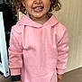 child, smiling, curly_hair, pink_hoodie, polka_dot_leggings, indoor, door, floor, person, happy, standing, casual_clothing, young_child, face, feet, shoes, wooden_door, background, home, expression
