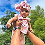 baby, child, pink_bow, headwrap, floral_top, hands, adult, outdoor, blue_sky, greenery, watch, portrait, cute, smile, person, nature, daylight, holding, happy, infant