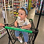 toddler, child, shopping_cart, pacifier, headband, blue_pants, white_shoes, bracelet, store_aisle, candy, snack_zone, shopping, retail, floor, hand, person, clothing, indoor, grocery, curious