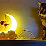 animal, cat, crescent_moon, curious, decor, ears, eyes, figurine, glowing, indoor, lamp, light, nighttime, pet, shadow, shelf, tabby_cat, toy, whiskers, wood