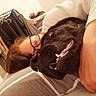 person, dog, black_dog, glasses, cuddling, indoor, bed, pillow, clothing_rack, smiling_dog, teeth, face, hair, hand, sleeve, blanket, casual, cozy, home, relaxing