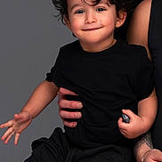 Gabriel a rejoint le concours — aidez-le/la à gagner de superbes lots ! toddler, child, curly_hair, black_clothing, smiling, playful, portrait, studio, adult_hand, holding, neutral_background, happy, cute, person, indoors, short_sleeves, casual_wear, young_child, human, expression