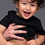 Gabriel participe au concours pour gagner de l'argent avec cette photo : toddler, child, curly_hair, smiling, laughing, black_shirt, belly, hand, adult_hand, playing, happy, cute, portrait, studio, gray_background, person, young_child, casual_clothing, expressive_face, joy