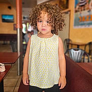Isabella a rejoint le concours — aidez-le/la à gagner de superbes lots ! black_shorts, blurry_background, brown_hair, cafe, chair, child, clothing, curious, curly_hair, expression, furniture, indoor, person, portrait, serious, skin, standing, table, toddler, yellow_top