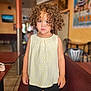 black_shorts, blurry_background, brown_hair, cafe, chair, child, clothing, curious, curly_hair, expression, furniture, indoor, person, portrait, serious, skin, standing, table, toddler, yellow_top