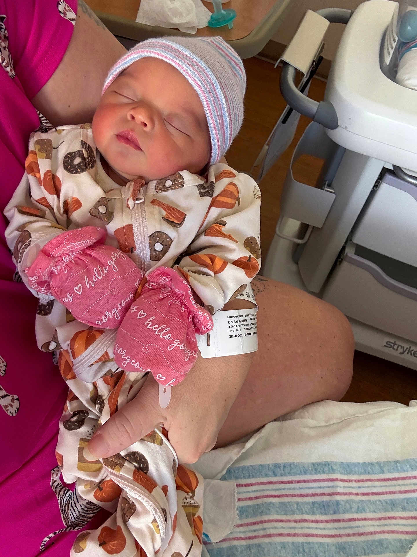 Kinsley is registered to the contest to win money with this photo: newborn, baby, infant, mittens, hat, onesie, sleeping, hospital, blanket, adult_hand, person, cradle, closeup, pacifier, medical_bracelet, bed, wood_floor, pink_clothing, patterned_clothing, resting