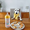 dog, puppy, white_dog, yellow_bib, sitting, table, dining_room, wooden_table, plate, pastries, bottle, baby_bottle, food, chair, lamp, houseplant, wall_art, indoors, portrait, funny