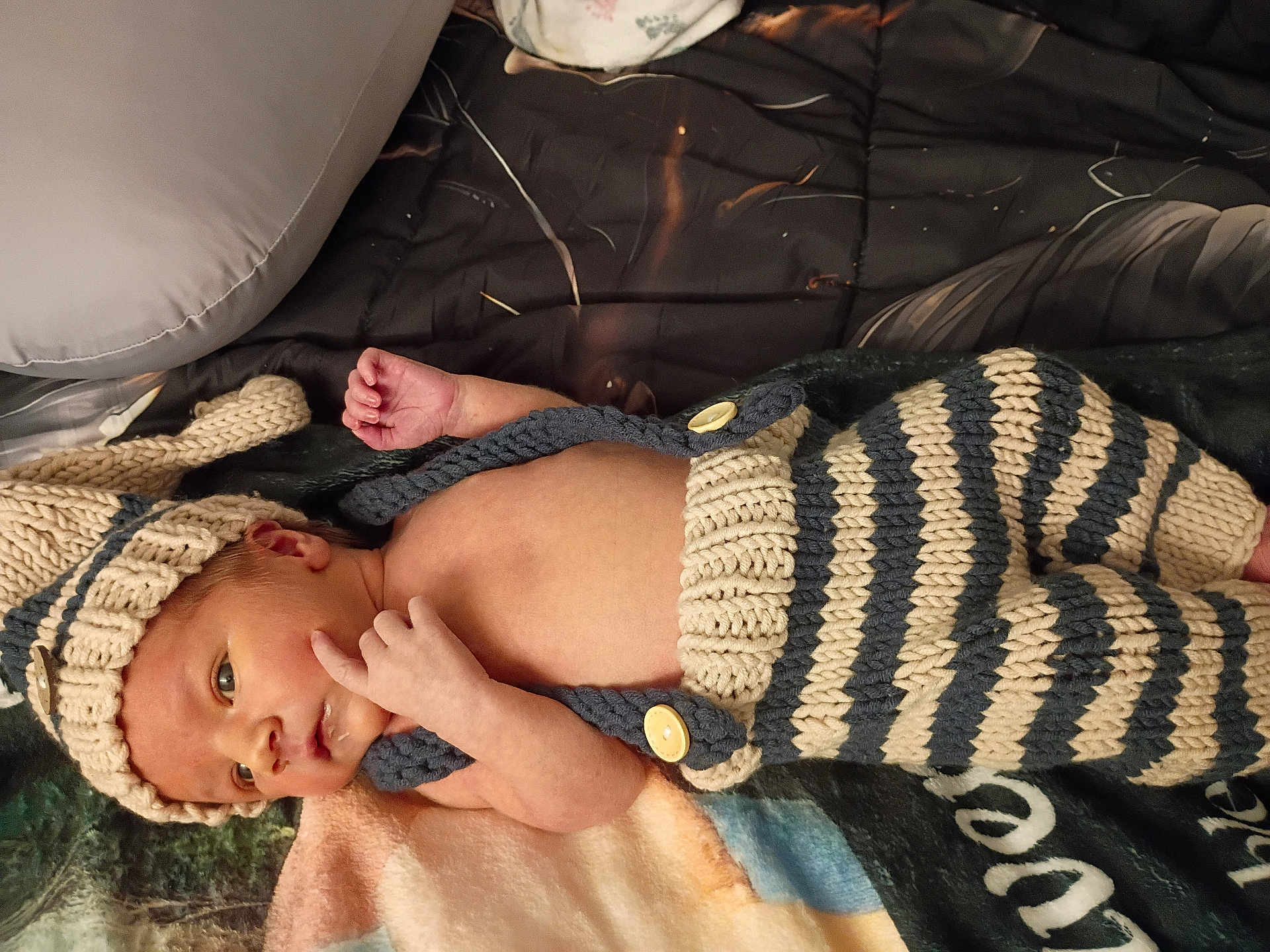 Colter is registered to the contest to win money with this photo: baby, newborn, infant, knitted_hat, knitted_overall, striped_pants, blanket, bedding, portrait, close_up, cozy, button, hand, face, awake, skin, soft_texture, indoor, nursery, adorable