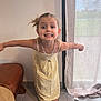 child, girl, smiling, yellow_dress, sandals, indoor, curtain, floor, tile_floor, necklace, playful, happy, posing, natural_light, window, furniture, brown_chair, person, standing, hair_tied