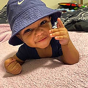 King is registered to the contest to win money with this photo: baby, child, smile, hat, bucket_hat, hand_gesture, blanket, pink_blanket, indoor, toy, black_and_white_blanket, clothing, face, person, cute, infant, lying_down, happy, portrait, headwear