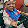 King joined the competition — help win amazing prizes! baby, smiling, hat, bib, high_chair, indoor, child, seated, happy, clothing, furniture, red_bench, table, person, cute, young_child, infant, portrait, baby_bib, baby_hat