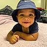 King is registered to the contest to win money with this photo: baby, child, smiling, face, hat, bucket_hat, blue_hat, blanket, pink_blanket, indoors, portrait, person, cute, happy, young_child, infant, lying_down, hand, skin, clothing