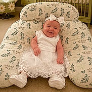 Ana Olivia is registered to the contest to win money with this photo: baby, smiling, white_dress, headband, bow, cushion, floral_pattern, carpet, toy, nursery, child, happy, seated, indoor, playful, cute, infant, decor, furniture, comfortable