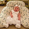 baby, smiling, white_dress, headband, bow, cushion, floral_pattern, carpet, toy, nursery, child, happy, seated, indoor, playful, cute, infant, decor, furniture, comfortable