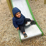 Kassym a rejoint le concours — aidez-le/la à gagner de superbes lots ! child, toddler, slide, playground, gravel, hooded_jacket, puffy_jacket, sneakers, smile, happy, playing, outdoor, park, metal_slide, green_trim, sitting, single_person, face, childhood, fun