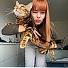 bed, bengal_cat, brown_sweater, casual, cat, dresser, furniture, green_eyes, holding, home, indoor, long_hair, person, pet, playful, portrait, red_hair, white_pants, winking, woman