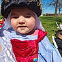 baby, child, costume, hat, necklace, red_sash, outdoor, park, grass, blue_sky, people, daylight, smiling, face, portrait, festive, celebration, clothing, person, event