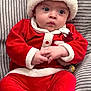 baby, infant, child, santa_hat, red_suit, white_fur_trim, velvet, couch, striped_cushion, wide_eyes, hands_clasped, sitting, holiday_outfit, festive, portrait, soft_texture, plush, indoor, cute, gaze
