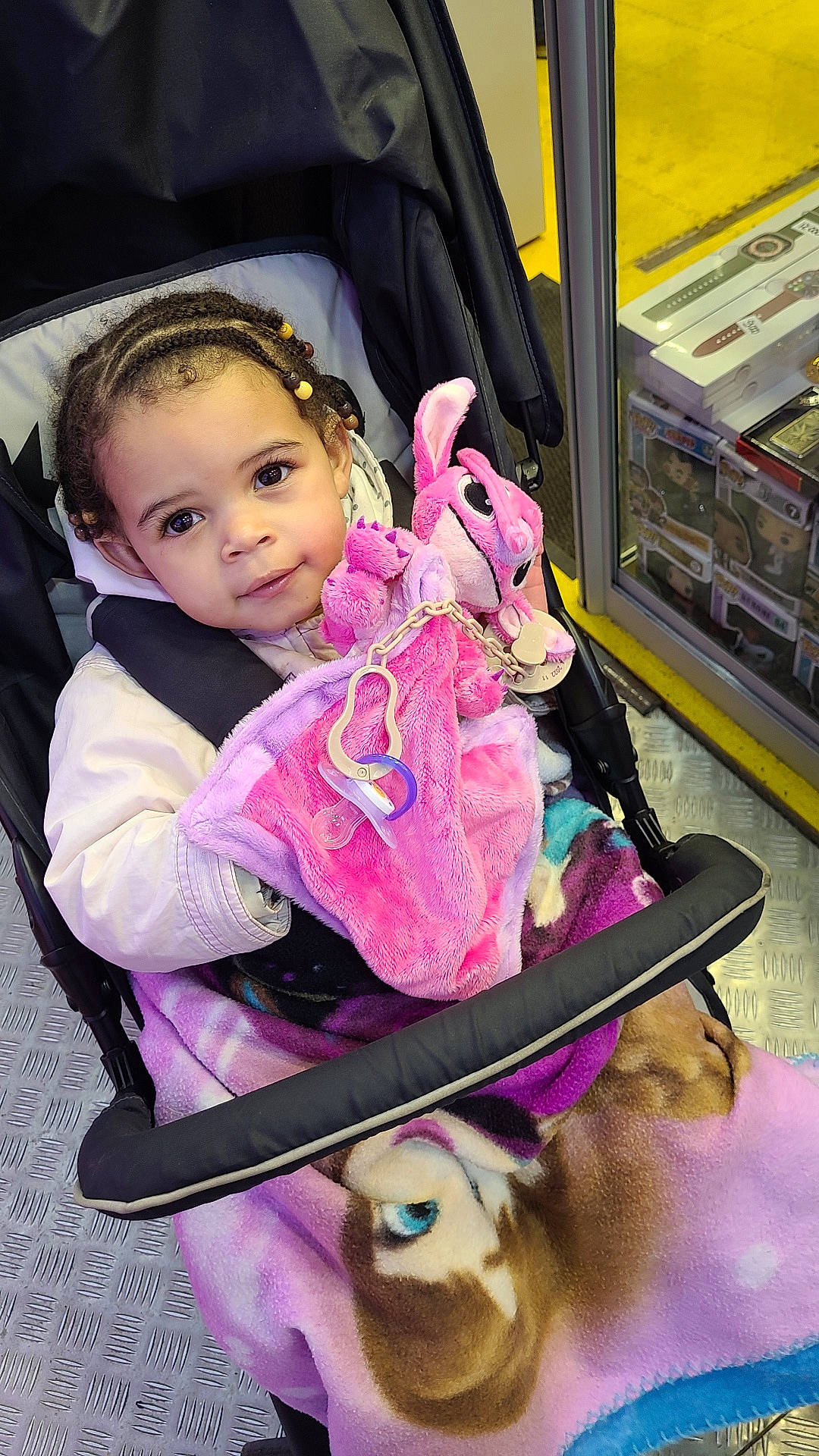 Hawa participe au concours pour gagner de l'argent avec cette photo : baby, blanket, braided_hair, child, clothing, colorful, cute, floor, indoor, person, plush_toy, portrait, retail, seated, shopping_area, smiling, stroller, toddler, toy, window_display