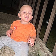 Anthony is registered to the contest to win money with this photo: toddler, child, sitting, stairs, outdoor, night, smiling, orange_shirt, gray_pants, concrete, wooden_railing, grass, happy, casual_clothing, person, young_child, steps, playful, small, cute