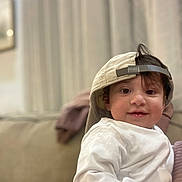Victoria Grace is registered to the contest to win money with this photo: toddler, child, smile, face, cap, white_shirt, indoor, couch, curtains, background_blur, person, hair, headwear, seated, young_child, happy, portrait, cute, casual, relaxed