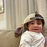 toddler, child, smile, face, cap, white_shirt, indoor, couch, curtains, background_blur, person, hair, headwear, seated, young_child, happy, portrait, cute, casual, relaxed