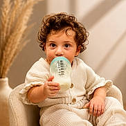 Victoria Grace is registered to the contest to win money with this photo: toddler, child, milk_bottle, curly_hair, cream_outfit, armchair, indoor, cozy, soft_lighting, neutral_background, baby, sitting, cute, portrait, curious, hand, face, eyes, furniture, warm_colors