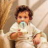 toddler, child, milk_bottle, curly_hair, cream_outfit, armchair, indoor, cozy, soft_lighting, neutral_background, baby, sitting, cute, portrait, curious, hand, face, eyes, furniture, warm_colors