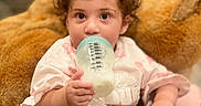 Victoria Grace joined the competition — help win amazing prizes! toddler, child, baby_bottle, milk, curly_hair, teddy_bear, toy, pink_clothing, indoor, person, cute, face, hand, sitting, soft_focus, cozy, background_blur, young_child, feeding, infant