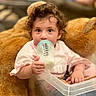 toddler, child, baby_bottle, milk, curly_hair, teddy_bear, toy, pink_clothing, indoor, person, cute, face, hand, sitting, soft_focus, cozy, background_blur, young_child, feeding, infant