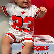 Ezel is registered to the contest to win money with this photo: baby, child, clothing, cute, diapers_box, footwear, happy, indoor, number_23, person, playful, red_background, shorts, sitting, smiling, toddler, tshirt, white_background, white_shoes, young_child