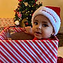Ezel is registered to the contest to win money with this photo: baby, celebration, child, christmas_hat, christmas_tree, cute, decorations, face, festive, gift_box, holiday, holiday_lights, indoors, ornaments, present, red_and_white, seasonal, sitting, smiling, striped_wrapping_paper