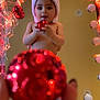 blurred_foreground, celebration, child, christmas_ornament, cute, decorations, face, festive, hand, holiday, indoors, joy, mirror, red, reflection, santa_hat, smiling, string_lights, toddler, warm_lighting