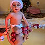 Ezel is registered to the contest to win money with this photo: baby, celebration, child, christmas_ornaments, christmas_tree, cute, decorations, diaper, face, festive, floor, holiday, indoor, lighting, presents, red_socks, reflection, santa_hat, smiling, toddler