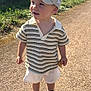 toddler, child, boy, cap, hat, striped_shirt, shorts, sneakers, outdoor, sunlight, gravel_path, shadow, snack, summer, casual_clothing, daytime, greenery, fence, house, curious_expression