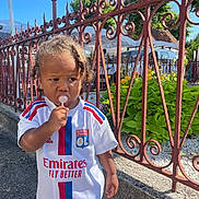 Sélas a rejoint le concours — aidez-le/la à gagner de superbes lots ! casual_clothing, child, concrete, curly_hair, daylight, expression, greenery, iron_fence, lollipop, olympique_lyonnais, outdoor, plants, shorts, sideways_gaze, sports_jersey, street, summer, sunny, toddler, young_child