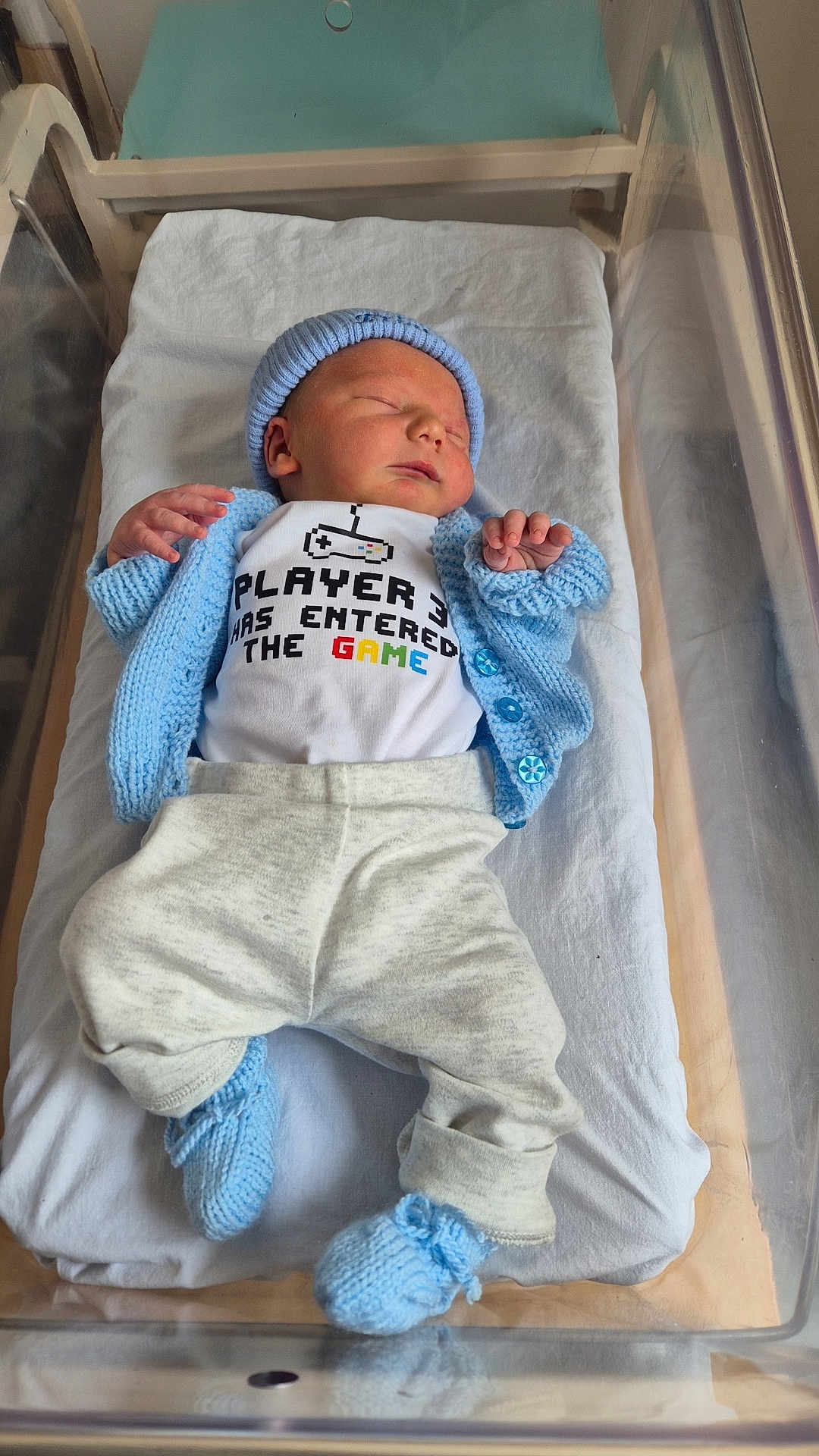 Dante is registered to the contest to win money with this photo: newborn, baby, sleeping, hospital, bassinet, blue_hat, blue_sweater, blue_booties, white_onesie, text_on_clothing, gray_pants, infant, bed, blanket, indoor, person, cute, sleep, child, knitwear