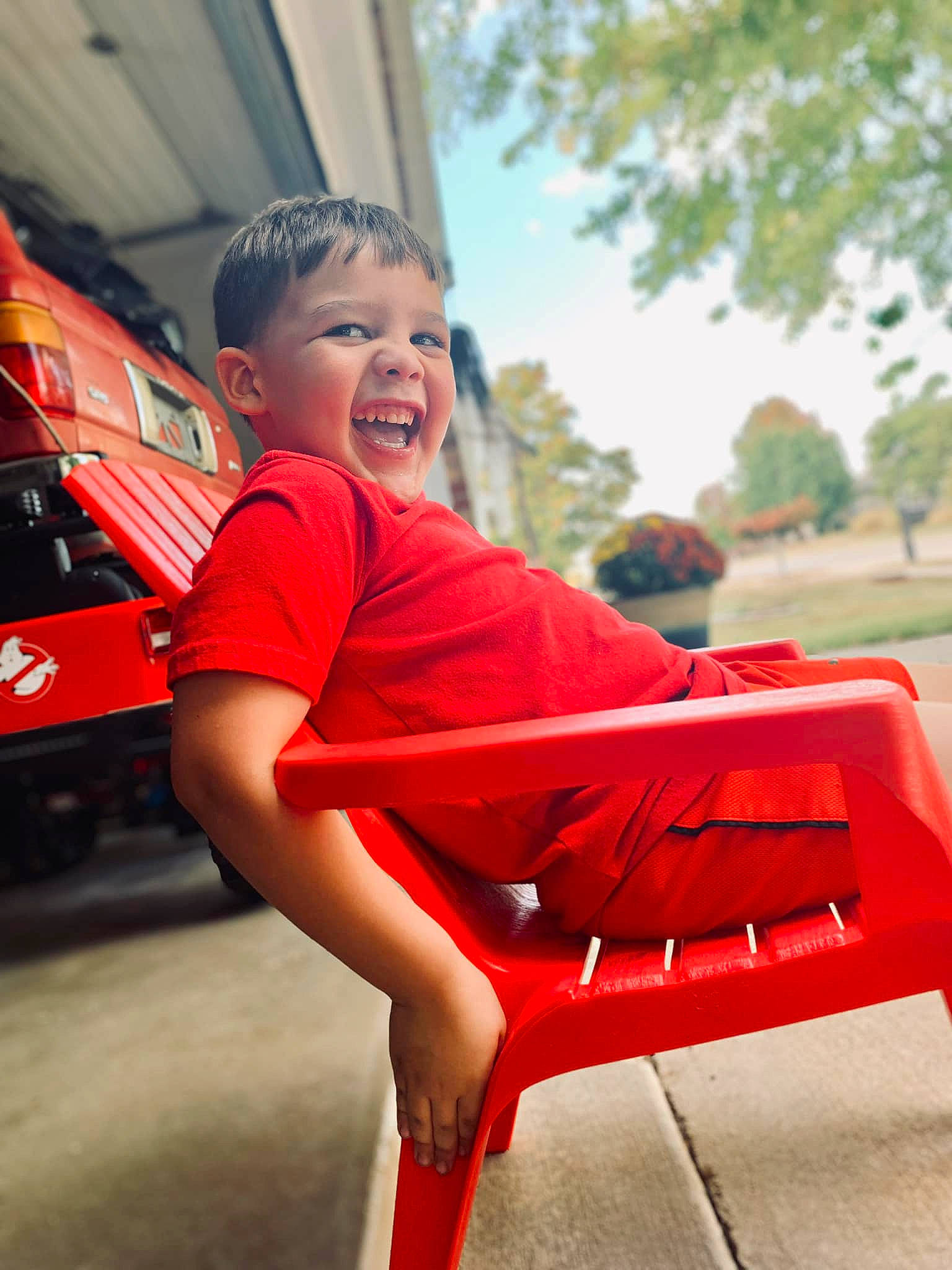 Ryker is registered to the contest to win money with this photo: automotive_design, automotive_exterior, automotive_lighting, bumper, fun, grass, happy, human_leg, leisure, motor_vehicle, orange, person, recreation, red, shorts, sky, smile, t_shirt, toddler, tree