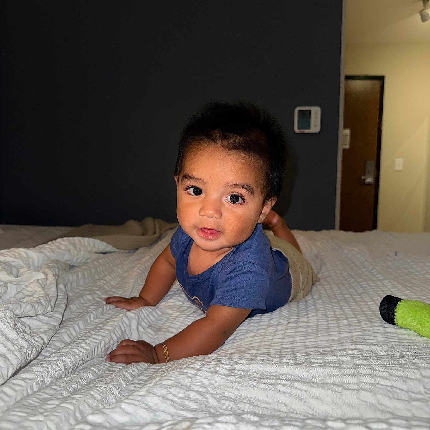 Norlan is registered to the contest to win money with this photo: baby, child, infant, portrait, indoors, bedroom, bed, blanket, blue_shirt, sitting, crawling, smiling, curious, dark_hair, face, eyes, hands, thermostat, door, toy