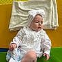 baby, child, white_clothing, headband, bow, blanket, colorful_background, mat, sitting, infant, cute, indoors, soft_texture, sock, footwear, portrait, young_child, expression, hand, person